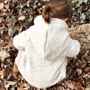 May include: A young child wearing a light beige hooded sweater sits on a bed of brown leaves. The child's hair is pulled back in a ponytail.