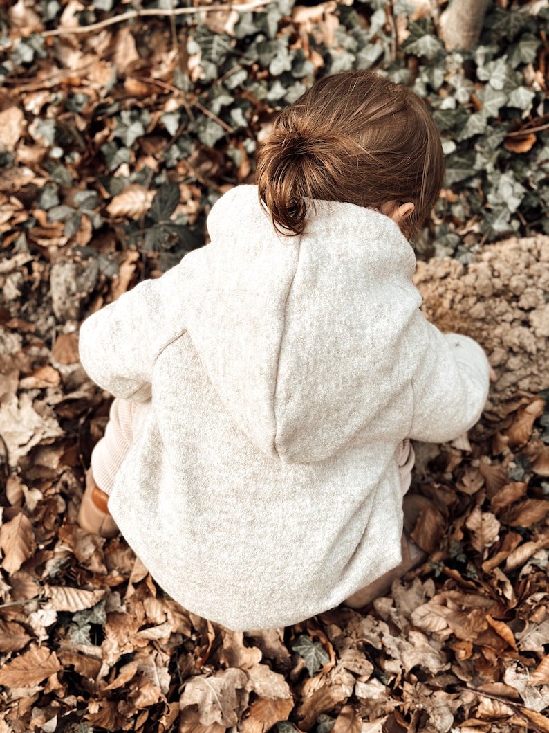May include: A young child wearing a light beige hooded sweater sits on a bed of brown leaves. The child's hair is pulled back in a ponytail.