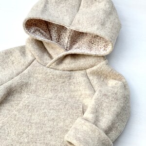 May include: A beige hooded sweatshirt with a brown leopard print lining. The sweatshirt has a pocket on the front and long sleeves with cuffed ends.