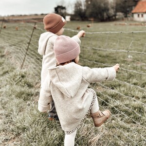 May include: Two children wearing brown and pink knit hats look at a fence with a grassy field behind them.