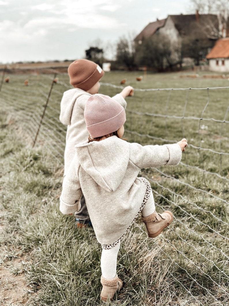 May include: Two children wearing brown and pink knit hats look at a fence with a grassy field behind them.