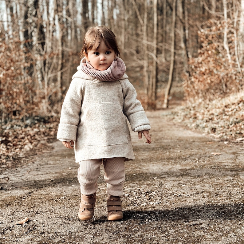May include: A young child wearing a beige sweater with a pink cowl neck scarf and brown boots walks on a path in a forest.