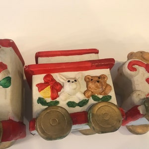 May include: A vintage ceramic Christmas train set with four cars. The first car features Santa Claus with the word "Happy" on the side. The second car has a white bunny, a brown bear, and a red bow with a bell. The third car has a brown bear holding a candy cane. The fourth car has a white cat with a red bow. All cars have gold wheels.