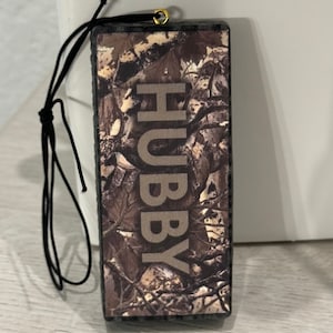 Peut inclure: Une décoration en bois de couleur camouflage marron et noir avec une ficelle noire pour l'accrocher. La décoration porte l'inscription "HUBBY" en blanc.