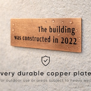Op de afbeelding: Een rechthoekige koperen plaat met de tekst "The building was constructed in 2022". De plaat is gemonteerd op een getextureerd grijs oppervlak. De tekst "very durable copper plate" staat eronder.