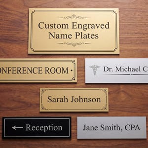 May include: A selection of custom engraved name plates. The plates feature text including "Conference Room," "Sarah Johnson," and "Reception." The plates are in gold, silver, and black, displayed on a wooden surface. Measurements are not included.