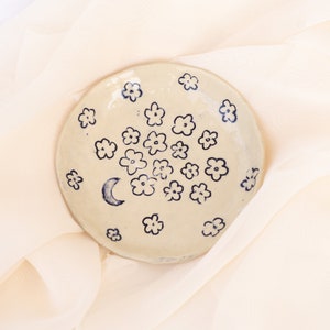 May include: A small, round, off-white ceramic dish decorated with hand-painted blue flowers and a crescent moon. The dish is placed on a soft, cream-colored fabric, creating a delicate and artistic presentation.