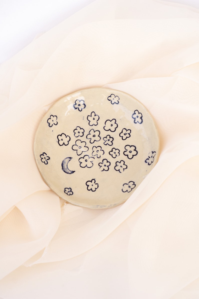 May include: A small, round, off-white ceramic dish decorated with hand-painted blue flowers and a crescent moon. The dish is placed on a soft, cream-colored fabric, creating a delicate and artistic presentation.