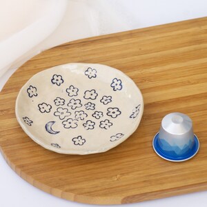 May include: A small, off-white ceramic dish decorated with blue floral and crescent moon designs. A silver and blue coffee pod sits next to the dish on a wooden cutting board. The dish is handmade and has a rustic, artisanal feel.