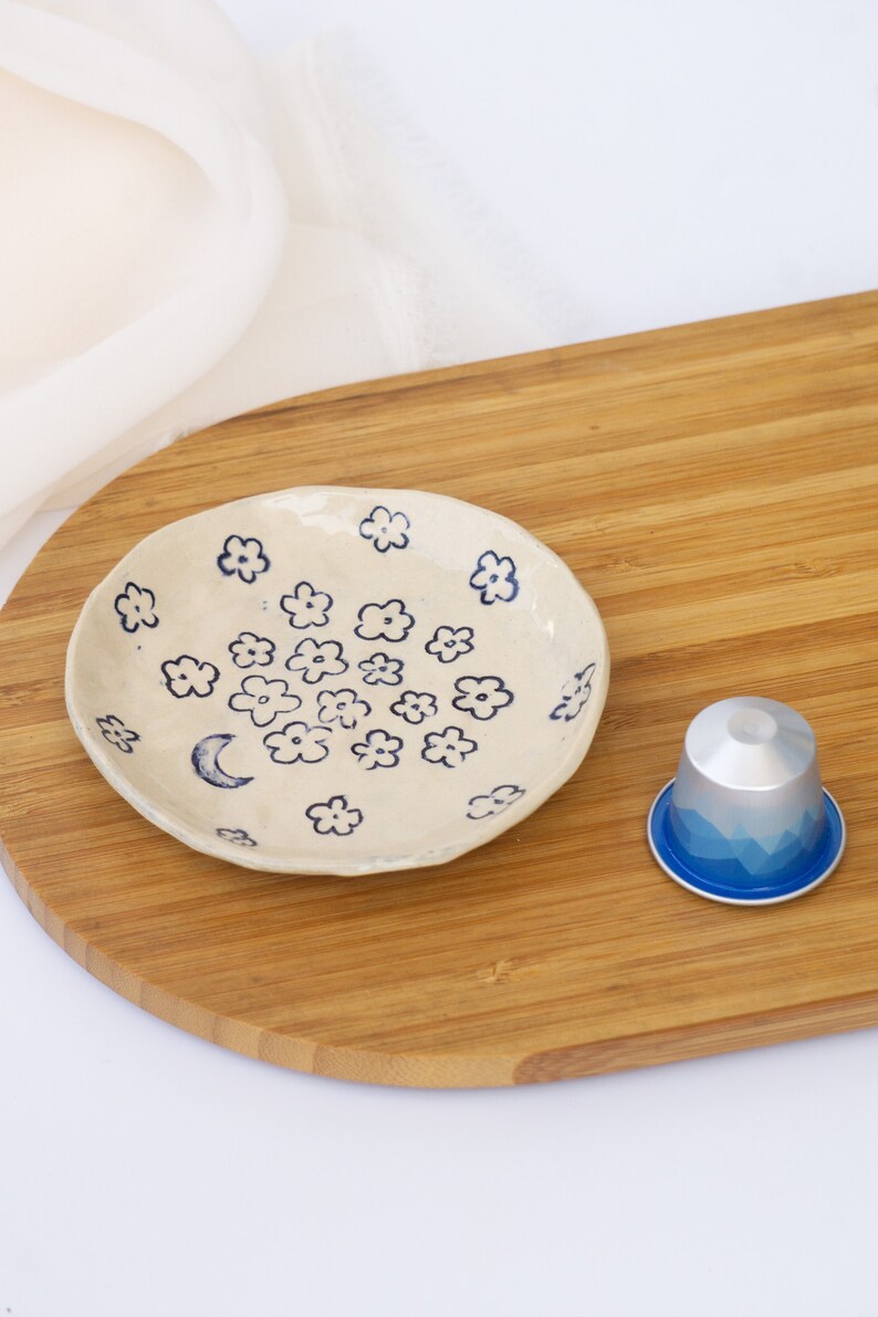 May include: A small, off-white ceramic dish decorated with blue floral and crescent moon designs. A silver and blue coffee pod sits next to the dish on a wooden cutting board. The dish is handmade and has a rustic, artisanal feel.
