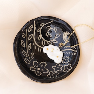 Pu&ograve; includere: Un vassoio portagioie in ceramica nera con un motivo floreale e a foglie, che contiene un ciondolo bianco a forma di nuvola con dettagli dorati e una catena dorata. Il piatto misura circa 10 cm di diametro.
