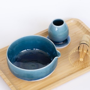 Puede incluir: Un cuenco de matcha de cerámica color turquesa con un pico, un jarrón pequeño y un batidor sobre una bandeja de bambú. El cuenco tiene un interior azul oscuro. El batidor es de bambú claro.
