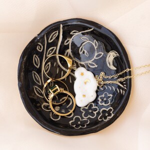 Pu&ograve; includere: Un vassoio portagioie in ceramica nera con un design floreale e lunare, contenente anelli d'oro e una collana con un ciondolo a forma di nuvola. Il piatto ha una superficie strutturata ed &egrave; decorato con dettagli bianchi e dorati.