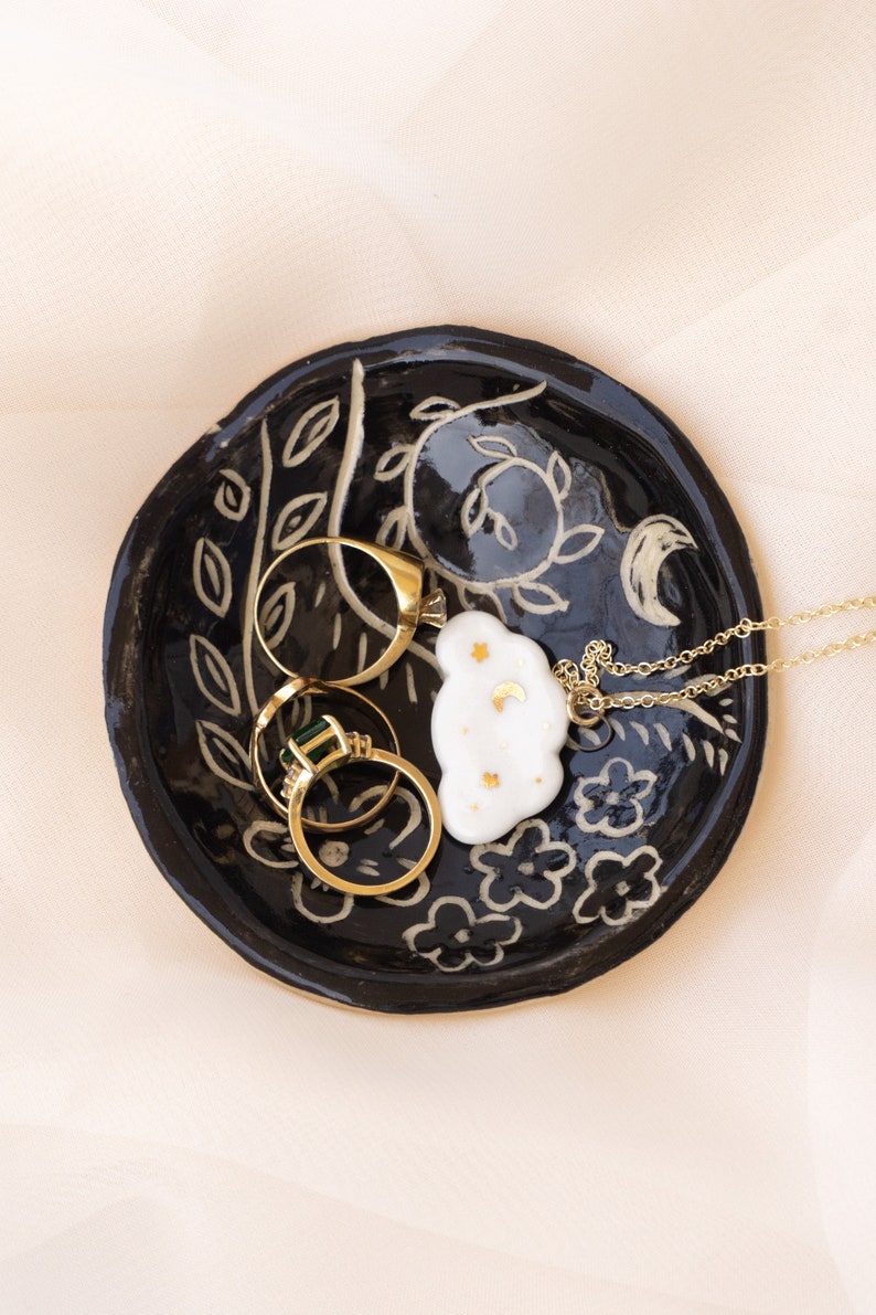 Pu&ograve; includere: Un vassoio portagioie in ceramica nera con un design floreale e lunare, contenente anelli d'oro e una collana con un ciondolo a forma di nuvola. Il piatto ha una superficie strutturata ed &egrave; decorato con dettagli bianchi e dorati.