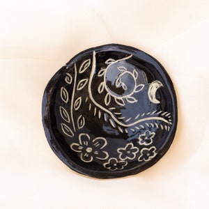 Pu&ograve; includere: Un piccolo piatto rotondo in ceramica nera lucida. Decorato con disegni floreali e di foglie bianche. Il bordo &egrave; leggermente rialzato e la forma generale &egrave; organica. Il piatto &egrave; probabilmente fatto a mano.