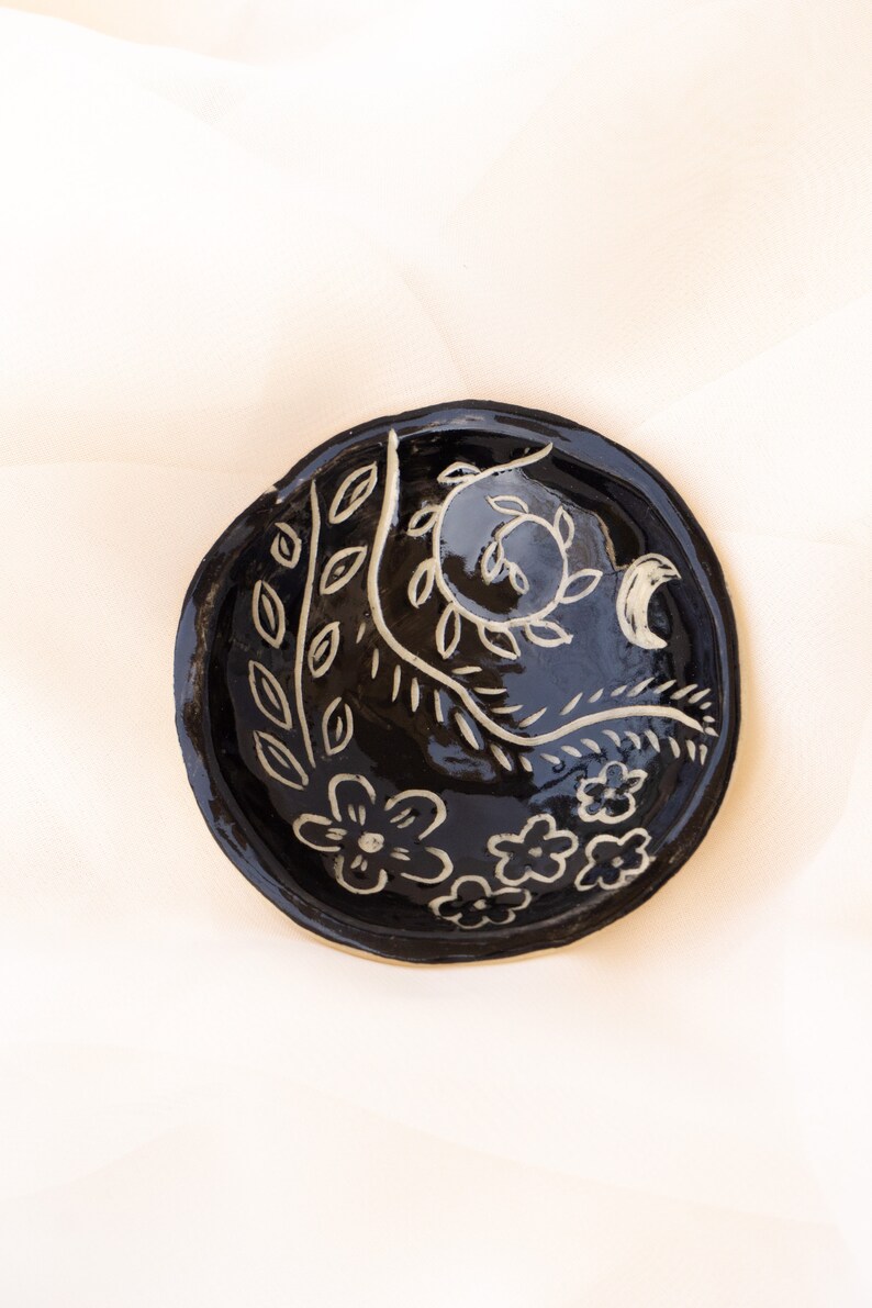 Pu&ograve; includere: Un piccolo piatto rotondo in ceramica nera lucida. Decorato con disegni floreali e di foglie bianche. Il bordo &egrave; leggermente rialzato e la forma generale &egrave; organica. Il piatto &egrave; probabilmente fatto a mano.