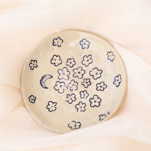 May include: A round, off-white ceramic dish decorated with dark blue floral designs and a crescent moon. The flowers are scattered across the surface. The dish is displayed on a soft, cream-colored fabric.