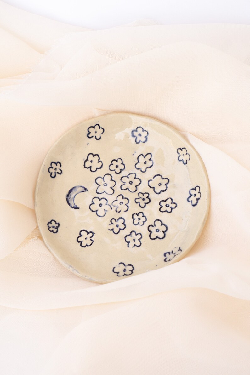 May include: A round, off-white ceramic dish decorated with dark blue floral designs and a crescent moon. The flowers are scattered across the surface. The dish is displayed on a soft, cream-colored fabric.