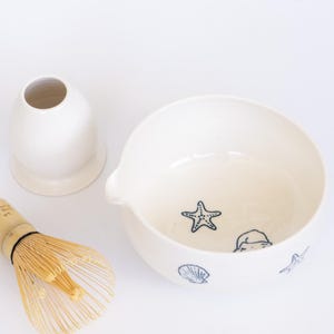 Puede incluir: Un cuenco de matcha de cerámica blanca con boquilla, decorado con ilustraciones azules de conchas marinas y estrellas de mar. También hay un batidor de bambú y un jarrón de cerámica blanca, creando un tema costero.