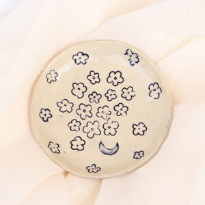 May include: A handmade, off-white ceramic plate with a hand-painted floral design in navy blue. The plate features a crescent moon and numerous small flowers. The plate is round and has a slightly uneven edge.