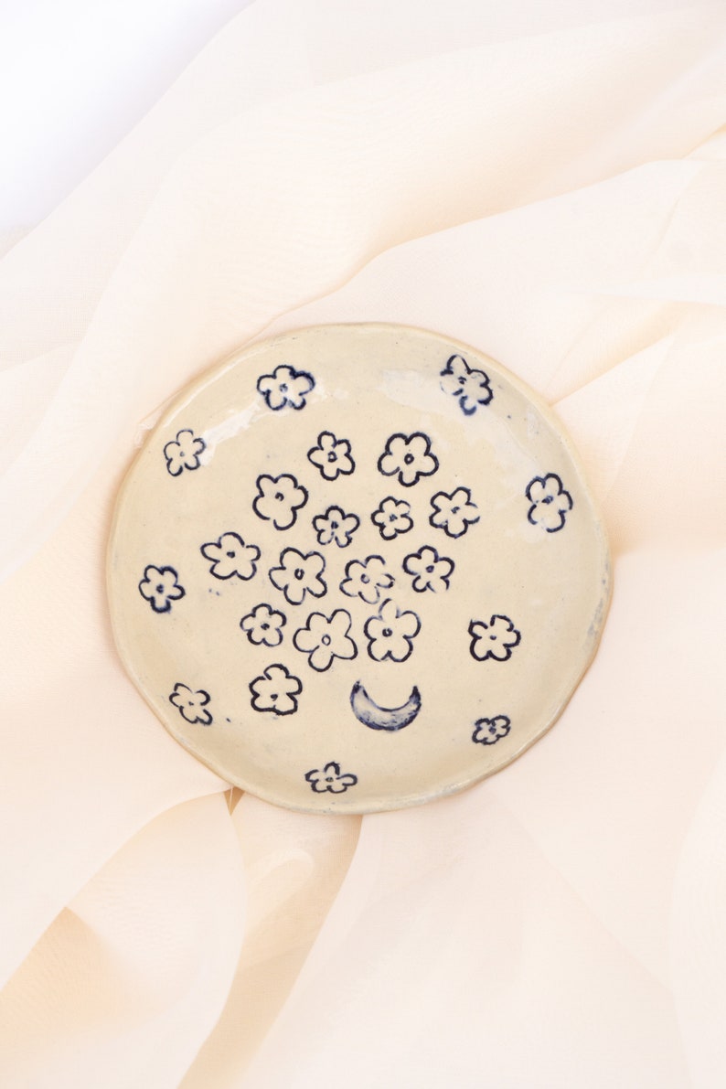 May include: A handmade, off-white ceramic plate with a hand-painted floral design in navy blue. The plate features a crescent moon and numerous small flowers. The plate is round and has a slightly uneven edge.