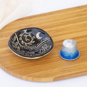 Pu&ograve; includere: Un piccolo piatto in ceramica nera con un motivo floreale bianco e una luna crescente. Accanto al piatto c'&egrave; una capsula di caff&egrave; argento e blu. Gli oggetti sono su un tagliere di legno.