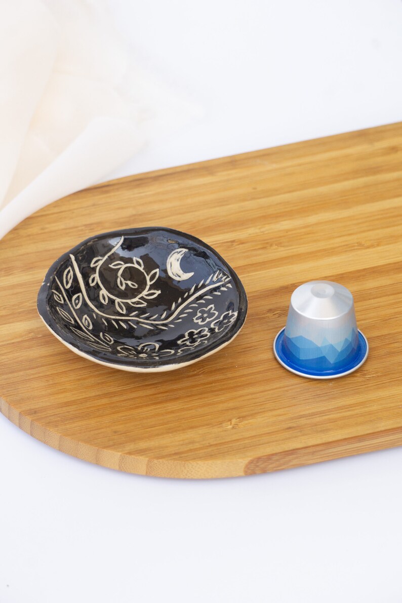 Pu&ograve; includere: Un piccolo piatto in ceramica nera con un motivo floreale bianco e una luna crescente. Accanto al piatto c'&egrave; una capsula di caff&egrave; argento e blu. Gli oggetti sono su un tagliere di legno.
