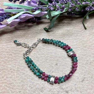 May include: A beaded bracelet featuring alternating rows of emerald green and ruby red gemstones. The bracelet has a silver-toned chain and decorative metal accents. The gemstones are small and faceted, creating a sparkling effect. The bracelet is displayed on a neutral surface.