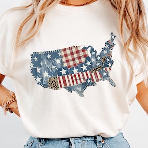 May include: Cream-colored t-shirt featuring a patchwork design of the United States map. The map incorporates denim, red and white stripes, stars, and leopard print patterns, creating a patriotic and unique graphic.
