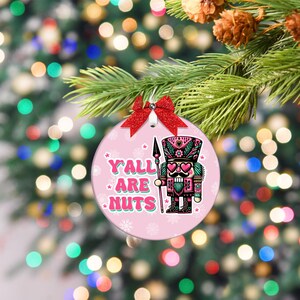 May include: A pink Christmas bauble with a red bow and a silver bell. The bauble features a colourful illustration of a nutcracker soldier with the text "Y'ALL ARE NUTS". The bauble is hanging on a Christmas tree.
