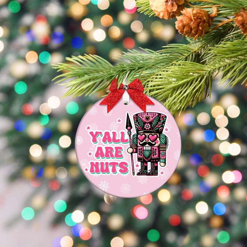 May include: A pink Christmas bauble with a red bow and a silver bell. The bauble features a colourful illustration of a nutcracker soldier with the text "Y'ALL ARE NUTS". The bauble is hanging on a Christmas tree.