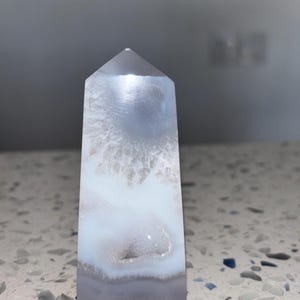 Puede incluir: Una punta de cristal de ágata blanca y gris con una parte superior puntiaguda y una superficie lisa y pulida. El cristal tiene un patrón único de bandas blancas y grises.