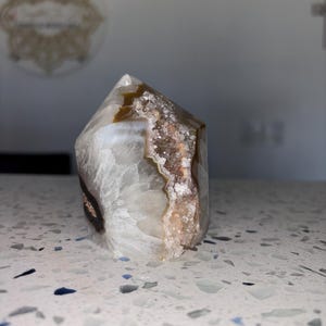Puede incluir: Una punta de cristal blanca, marrón y rosa con una textura natural, cruda y rugosa. La punta de cristal es una formación natural con una forma y un patrón únicos.