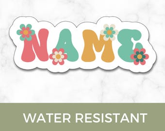 May include: A white sticker with the word "NAME" in colorful, bubble letters. The letters are decorated with flowers. The sticker also has the text "WATER RESISTANT STICKER".