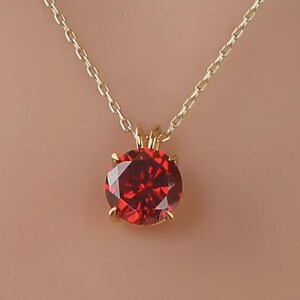 May include: A gold-coloured necklace featuring a round, faceted, red gemstone pendant. The pendant is held by four prongs. The chain has a simple link design. The background is a solid, light brown colour.