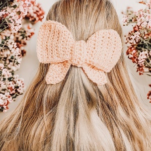 May include: A light pink crocheted bow hair clip. The bow is tied in a classic shape with two loops and a center knot.
