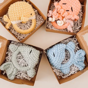 Peut inclure: Quatre hochets de bébé en crochet dans des boîtes en carton avec des couvercles transparents. Les hochets ont la forme d'une baleine jaune, d'une pieuvre rose, d'un poisson bleu clair et d'un poisson bleu. Chaque hochet a un anneau et est entouré de papier déchiqueté.
