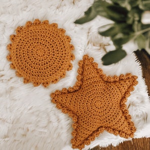May include: Two handmade crochet cushions in a warm, golden-brown hue. One is round with a scalloped edge, and the other is star-shaped, also with a scalloped border. The cushions are placed on a fluffy, white surface, creating a cosy, textured look.