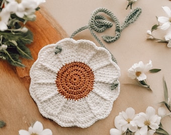 May include: A white crocheted daisy flower bag with a brown center and a green strap. The bag is surrounded by white flowers.