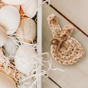 May include: A crocheted bunny rabbit ornament in a light brown color. The bunny is sitting on a white wooden surface with a basket of eggs in the background.