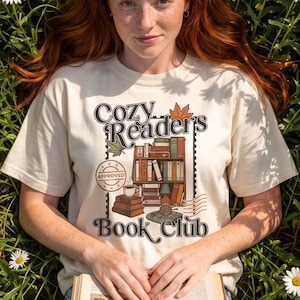 Op de afbeelding: Een crèmekleurig t-shirt met een grafische print van een boekenplank, een stapel boeken en een koffiekopje met de tekst "Cozy Reader's Book Club" en "Approved" in een stempelontwerp. Het shirt wordt gedragen met een lichtblauwe spijkerbroek.