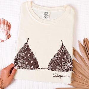 Puede incluir: Una camiseta de manga corta color crema con un gráfico marrón oscuro de una parte superior de bikini con estampado de bandana paisley. El texto "Estefania" está impreso en letra cursiva debajo del gráfico. La etiqueta interior dice "Comfort Colors".