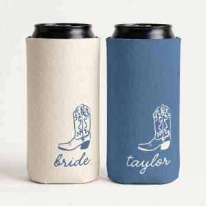 May include: Two slim can coolers with black rims. The left cooler is cream-colored with a blue cowboy boot graphic and the word bride in blue script. The right cooler is blue with a white cowboy boot graphic and the word taylor in white script.