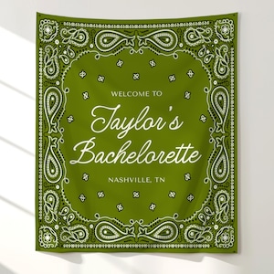 May include: A rectangular olive green fabric wall hanging featuring a white paisley bandana border pattern. The center text reads "WELCOME TO Taylor's Bachelorette NASHVILLE, TN" in a combination of white serif and script typography.