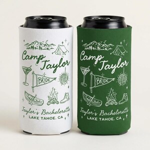 May include: Two tall beverage can coolers, one white and one dark green, each featuring a dark green graphic print. The design includes mountains, a tent, a martini glass, a pennant flag labeled BRIDE, a disco ball, a canoe, a campfire, and a boot. Text reads Camp Taylor, Taylor's Bachelorette, and LAKE TAHOE, CA.
