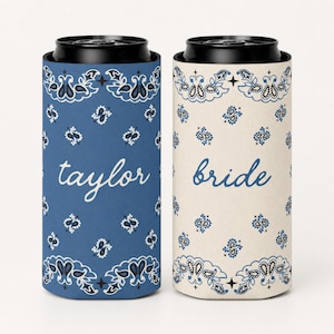 May include: Two cylindrical can coolers with black rims. The left cooler is blue with a white paisley pattern and the text "taylor" in script. The right cooler is off-white with a blue paisley pattern and the text "bride" in script.