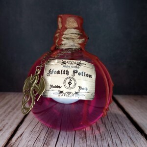May include: A decorative, pink glass bottle with a cork stopper and a label that reads "Health Potion Bubble Bath." A bronze dragon charm hangs from a chain around the bottle's neck. The bottle is on a wooden surface.
