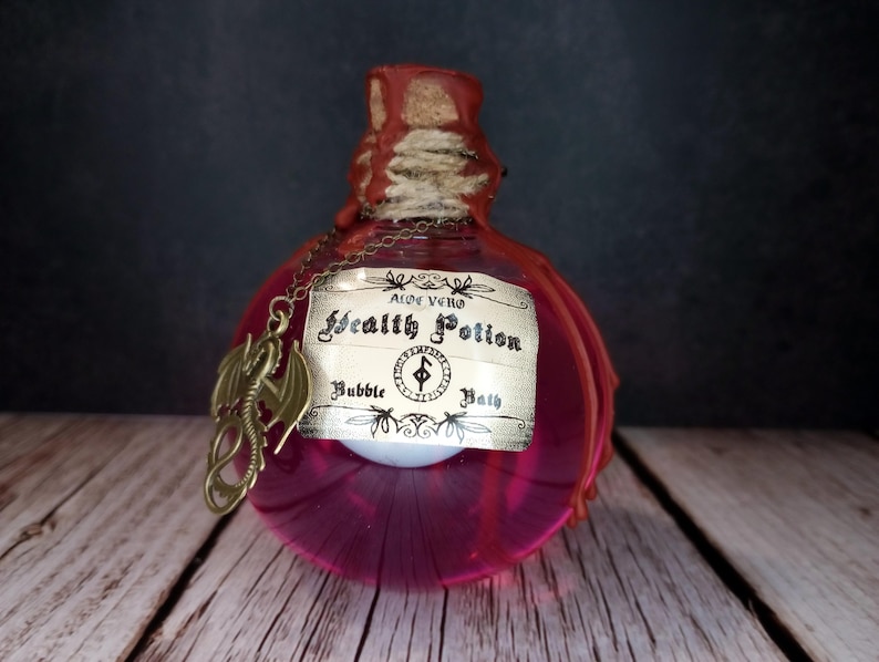 May include: A decorative, pink glass bottle with a cork stopper and a label that reads "Health Potion Bubble Bath." A bronze dragon charm hangs from a chain around the bottle's neck. The bottle is on a wooden surface.