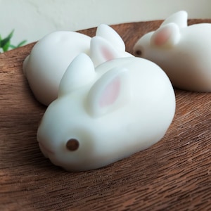 May include: Three white bunny-shaped soaps on a wooden surface. The soaps have pink ears and brown eyes.