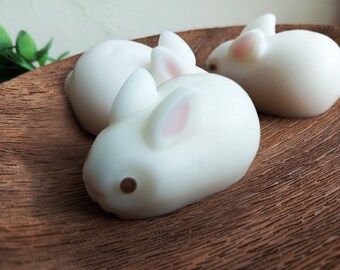May include: Three white bunny-shaped soaps on a wooden surface. The soaps have pink ears and brown eyes.